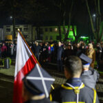 DSC_7832 Na zdjęciu widoczna uroczystość nocą z ludźmi i flagą Polski. Tło to oświetlone budynki i drzewa. W centrum stoi grupa ludzi w ciemnych ubraniach.