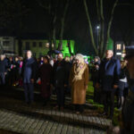 DSC_7838 Grupa ludzi stoi na bruku, w nocnej scenerii. Tło rozświetlają zielone światła budynków. Obecne są flagi i mundury.