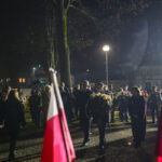 DSC_7850 Grupa osób składa wieńce pod pomnikiem nocą. Deszcz spada delikatnie, a w tle widać flagi Polski. Jasne lampy oświetlają kamienną alejkę.
