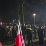 DSC_7854 Wieczorna uroczystość z tłumem ludzi i flagami Polski. Osoby niosą wieńce kwiatów, tło oświetlone latarniami. Pada lekki deszcz.