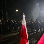 DSC_7860 Grupa ludzi w mundurach maszeruje nocą, trzymając kwiaty. Na pierwszym planie widoczne są flagi Polski. W tle lampa uliczna oświetla ciemne otoczenie.