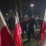 DSC_7861 Dwóch mężczyzn w mundurach niesie wieniec podczas nocnej uroczystości. Otaczają ich polskie flagi. W tle widoczne są postacie i latarnia uliczna.