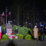 DSC_7866 Pomnik z popiersiem otoczony flagami Polski, wieńce kwiatowe z biało-czerwonymi wstęgami. W tle ciemny park, z przodu kobieta przy mikrofonie. Obok młodzieńcze harcerze w mundurach.