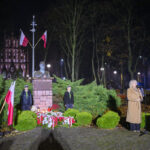 DSC_7867 Pomnik z popiersiem otoczony zielenią, udekorowany biało-czerwonymi flagami i kwiatami. Obok kobieta przemawia do mikrofonu. Nocne oświetlenie, w tle drzewa i budynek.