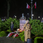 DSC_7870 Osoba w płaszczu przemawia przy mikrofonie przed pomnikiem, w tle flagi Polski. Zieleń roślin i nocne oświetlenie. Bukiety kwiatów u podstawy.