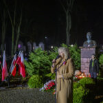 DSC_7873 Kobieta w płaszczu przemawia przed mikrofonem obok flag Polski. Tło to nocny park z pomnikiem i zielonymi krzewami. Widać także znicze i osoby w mundurach.