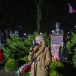 DSC_7875 Osoba przemawia nocą na tle pomnika i zielonych krzewów. Ubrana w beżowy płaszcz, trzyma mikrofon. W tle widać biało-czerwoną flagę.