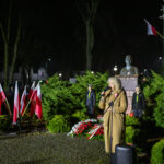 DSC_7877 Kobieta w płaszczu przemawia przy mikrofonie w parku. W tle są flagi polskie i pomnik. Nocna sceneria, kolorowe światło.