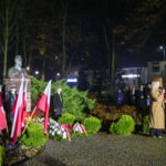 DSC_7882 Na zdjęciu jest popiersie przyozdobione flagami Polski w zielonym parku. Kobieta w płaszczu przemawia do mikrofonu. W tle grupa ludzi stoi nocą.