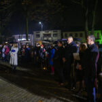 DSC_7889 Grupa ludzi w parku po zmroku, ubranych w ciemne płaszcze, otoczona drzewami. W tle budynki z zielonym oświetleniem.