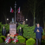 DSC_7898 Pomnik otoczony zielonymi krzewami, z flagami Polski nad popiersiem. Widoczne są wiązanki kwiatów w kolorach białym i czerwonym. Na pierwszym planie stoi mężczyzna przy mikrofonie.