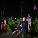 DSC_7899 W nocy, mężczyzna przemawia przy mikrofonie obok pomnika. W tle zielone krzewy i polskie flagi. Jasne oświetlenie i ciemne tło dodają kontrastu.