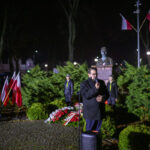 DSC_7901 Mężczyzna przemawia na tle zielonych krzewów nocą. Wokół niego stoją flagi Polski oraz wiązanki kwiatów. Za nim znajduje się pomnik i wartownicy.