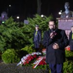 DSC_7903 Na zdjęciu mężczyzna przemawia przy mikrofonie, obok flagi Polski. W tle pomnik i zielone krzewy. Nocna atmosfera z oświetleniem ulicznym.