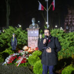 DSC_7904 Mężczyzna przemawia przy mikrofonie wieczorem, w tle pomnik z popiersiem, flagi i zielone krzewy. Uroczystość z kwiatami.