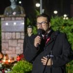 DSC_7905 Osoba w okularach przemawia do mikrofonu w parku nocą. W tle popiersie na cokole i zielone krzewy. Oświetlenie tworzy ciepły nastrój.