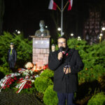 DSC_7908 Mężczyzna przemawia przy mikrofonie, obok pomnik z brązu i kwiaty, w tle polskie flagi na maszcie. Nocne oświetlenie rozjaśnia scenę. Ciemne tło z niebieskimi i czerwonymi akcentami.