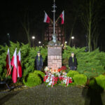 DSC_7910 Pomnik otoczony flagami polskimi, noc, oświetlony. Dwóch ludzi w mundurach stoi obok. Świece i kwiaty przed pomnikiem.