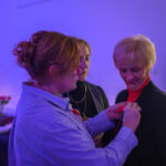 DSC_7919 Trzy kobiety w pomieszczeniu z ciepłym, fioletowym oświetleniem. Jedna przypina coś do ubrania innej. W tle stół z jedzeniem i świecami.