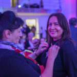 DSC_7921 Na zdjęciu dwie kobiety uśmiechają się w sali z fioletowym oświetleniem. Jedna przypina kokardkę do bluzki drugiej. W tle widać ludzi na czerwonych krzesłach.