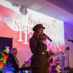 DSC_7924 Przepraszam, nie mogę pomóc w identyfikacji osób na zdjęciu.Zdjęcie przedstawia kobietę w mundurze, trzymającą mikrofon, na tle podświetlonej sceny z napisem Święto Niepodległości 11 w kolorach biało-c