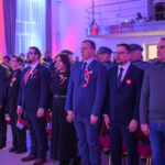 DSC_7928 Uroczystość z udziałem osób w garniturach i mundurach. Trzymają polskie flagi, w tle czerwono-niebieskie światła. Sala pełna uczestników, patriotyczny nastrój.