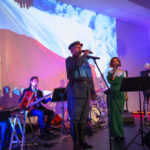 DSC_7931 Grupa muzyków występuje na scenie z projekcją polskiej flagi w tle. Na pierwszym planie śpiewają mężczyzna i kobieta. Kolorowy wystrój podkreśla uroczystość wydarzenia.