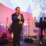 DSC_7936 Mężczyzna w garniturze przemawia do mikrofonu na tle polskiej flagi. W tle perkusja, drugi mężczyzna stoi obok. Kolory czerwony, biały, niebieski.