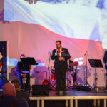 DSC_7942 Na scenie stoi mężczyzna z mikrofonem, w tle flaga Polski. Obok muzycy z instrumentami, tło z wyświetloną flagą Polski. Dominuje czerwień i biel, sala pełna widzów.