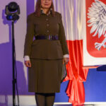 DSC_7947 Kobieta w brązowym mundurze wojskowym stoi na tle flagi Polski z godłem. Oświetlenie sceniczne w tle. Po lewej lampa sceniczna.