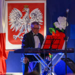 DSC_7948 Mężczyzna w smokingu gra na klawiszach na tle flagi z orłem w koronie. Obok kwiatu białego i czerwonego. Niebieskie tło sceny.