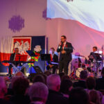 DSC_7949 Na scenie występują muzycy i wokalista, tło z polską flagą i orłem, dominują kolory biały i czerwony.