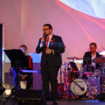 DSC_7951 Mężczyzna w eleganckim garniturze śpiewa na scenie. Towarzyszą mu muzycy przy instrumentach na tle polskiej flagi. Światła w kolorze czerwonym i niebieskim.