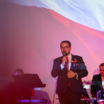 DSC_7956 Mężczyzna w garniturze śpiewa na scenie z mikrofonem. Tło w kolorach czerwonym i białym, z muzykiem grającym na perkusji.