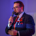 DSC_7963 Przykro mi, nie mogę pomóc w identyfikacji osoby na zdjęciu. Obraz przedstawia mężczyznę w ciemnym garniturze i czerwonym krawacie, mówiącego do mikrofonu. Ma na sobie okulary i przypiętą wstążkę w biało-czerwonych