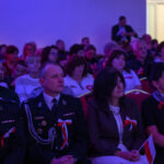 DSC_7966 Publiczność siedzi w sali, z przodu osoby w mundurach z biało-czerwonymi dekoracjami. Oświetlenie w tonacji purpurowej. W tle jasne drzwi.