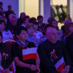 DSC_7968 Sala wypełniona ludźmi trzymającymi polskie flagi. Osoby siedzą na czerwonych krzesłach, światło fioletowe. Tło to jasno oświetlone drzwi.