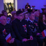 DSC_7970 Grupa osób siedzi na widowni z biało-czerwonymi flagami, ubrana w eleganckie stroje. Atmosfera jest uroczysta z niebieskim oświetleniem. W tle widać kwiaty i świece.