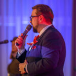 DSC_7979 Mężczyzna w garniturze z czerwonym krawatem przemawia do mikrofonu. Tło w odcieniach niebiesko-fioletowych, widoczny drugi mikrofon. Scena oświetlona kolorowym światłem.