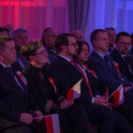 DSC_7982 Grupa ludzi siedzących w ciemnym pomieszczeniu z czerwonym i niebieskim oświetleniem. Osoby mają flagi Polski i biało-czerwone kotyliony. W tle są ciemne zasłony.