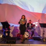 DSC_7991 Na scenie kobieta przemawia do mikrofonu, w tle flaga Polski. Obok zespół muzyczny, dominują czerwone i białe kolory. Tło ma patriotyczny charakter.