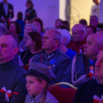 DSC_7998 Grupa ludzi w mundurach siedzi na widowni. Dominują niebieskie i czerwone kolory. W tle rozmyte, jasne światło.