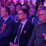 DSC_7999 Grupa ludzi siedzi na uroczystości, ozdobiona biało-czerwonymi kokardkami. Ubrani w eleganckie stroje. Tło niebiesko-różowe.