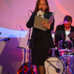 DSC_8003 Kobieta w czarnej sukni przemawia do mikrofonu, trzymając kartki. W tle widać perkusistę i kolorowe oświetlenie sceniczne.