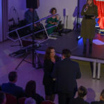 DSC_8015 Pomieszczenie z ludźmi na uroczystości. Kobieta na scenie w khaki przemawia, obok muzycy. Dominują kolory czerwony i niebieski.