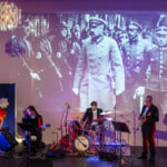 DSC_8025 Grupa muzyków gra na scenie z niebieskim oświetleniem. W tle duża, czarno-biała projekcja historycznych żołnierzy. Obok widać polskie godło na czerwonym tle.