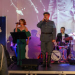 DSC_8028 Osoby śpiewające na scenie w eleganckich strojach, tło to biało-czarny obraz historyczny, zespół muzyczny z perkusją.