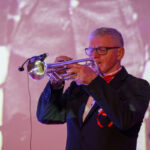 DSC_8031 Mężczyzna w garniturze gra na trąbce. Tło jest rozmyte, w odcieniach fioletu i różu. Muzyk nosi okulary i czerwony krawat.