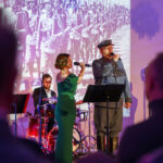 DSC_8034 Na scenie kobieta i mężczyzna śpiewają, z tyłu perkusista. Kobieta w zielonej sukni, mężczyzna w mundurze. Tło to czarno-białe zdjęcie żołnierzy.