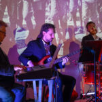 DSC_8035 Trzech muzyków gra: jeden na klawiszach, drugi na gitarze basowej, trzeci na perkusji. Tło z projekcją w sepii. Oświetlenie purpurowe i czerwone.